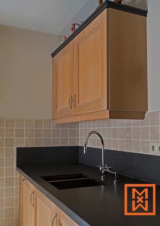 Moderne hoekkeuken met licht eikenhouten kasten en een strak zwart aanrechtblad met een dubbele spoelbak en een chromen kraan. De achterwand is van lichtbeige tegels en de kasten zijn versierd met decoratieve elementen.