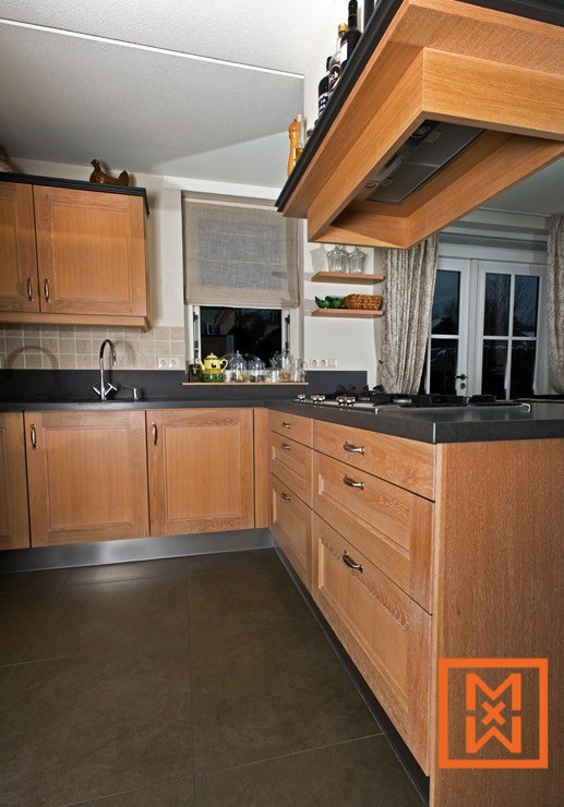 Modern keukenontwerp met licht eikenhouten kasten, donkere aanrechtbladen en een ingebouwde afzuigkap. De L-vormige keuken biedt veel opbergruimte en een strakke, minimalistische uitstraling. Een groot raam zorgt voor natuurlijk licht.