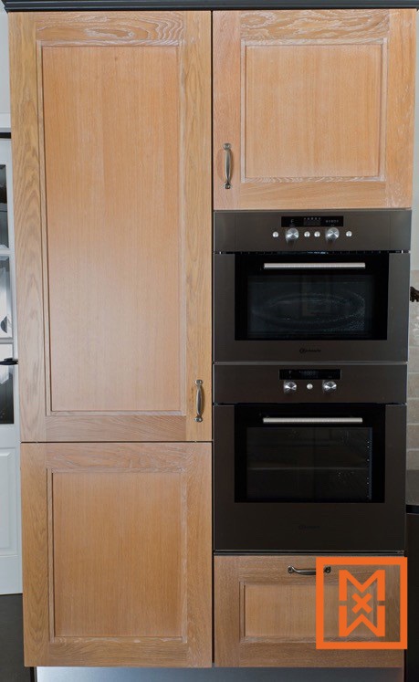 Licht eikenhouten keukenkastjes flankeren een ingebouwde, donkerbruine dubbele oven met daarboven een magnetron. De kasten zijn voorzien van paneeldeuren en eenvoudige, donkere metalen handgrepen. Onder de oven bevindt zich een kleine lade.