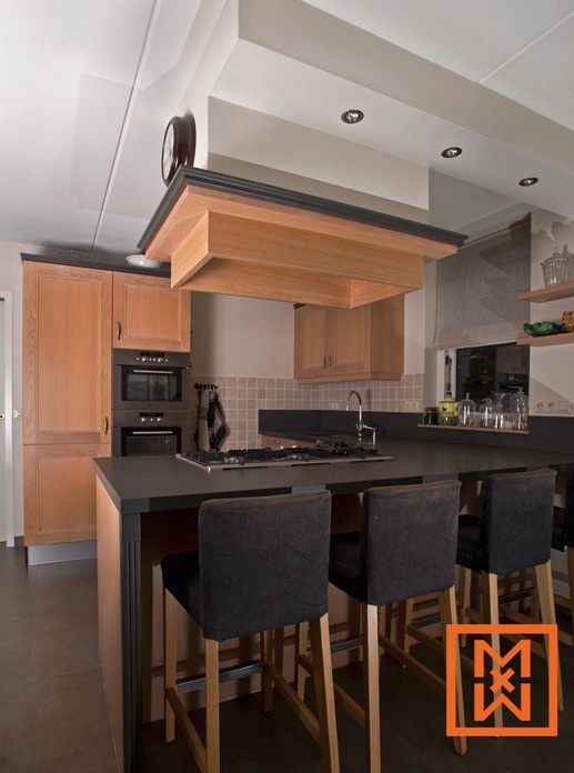 Modern keukenontwerp met een strak donker kookeiland met lichthouten barkrukken, verlicht door inbouwspots. De kasten zijn van licht hout, een mooie aanvulling op de donkere stenen werkbladen en creëren een warme en uitnodigende sfeer. Een op maat gemaakte afzuigkap van hout voegt een uniek architectonisch detail toe.