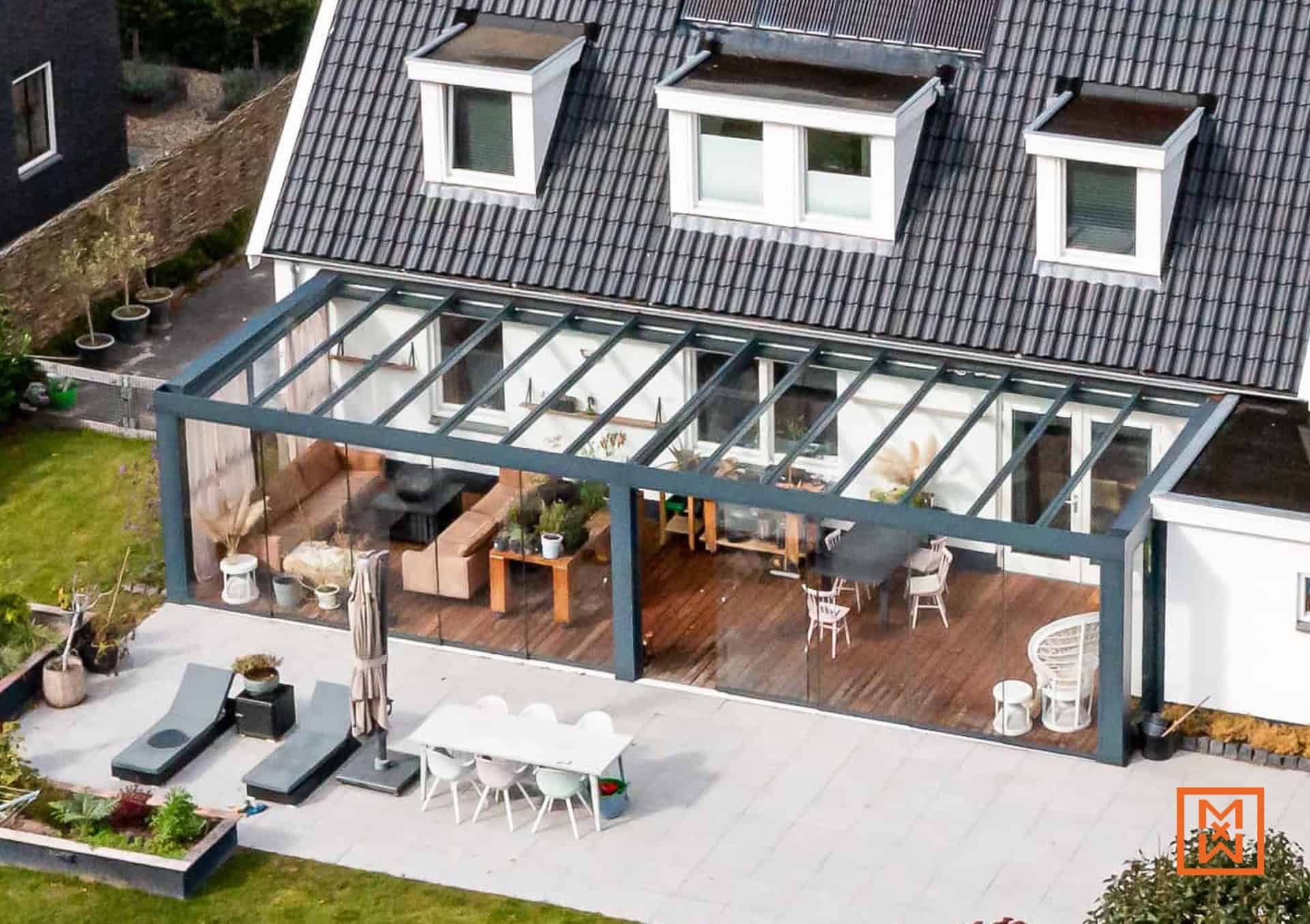 Moderne terrasoverkapping met glazen panelen en grijze metalen frames, grenzend aan een wit huis met donkergrijze dakpannen. Tuinmeubilair en beplanting creëren een gezellige buitenruimte.