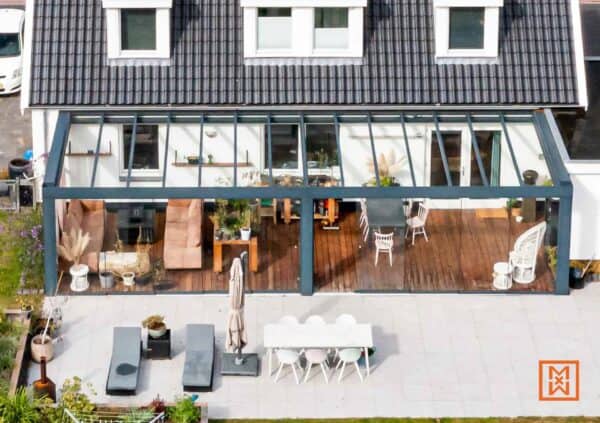 Een serre met een modern design, uitgerust met comfortabele meubels en een houten vloer, die een naadloze overgang biedt naar een betegeld terras met een eethoek en ligstoelen.