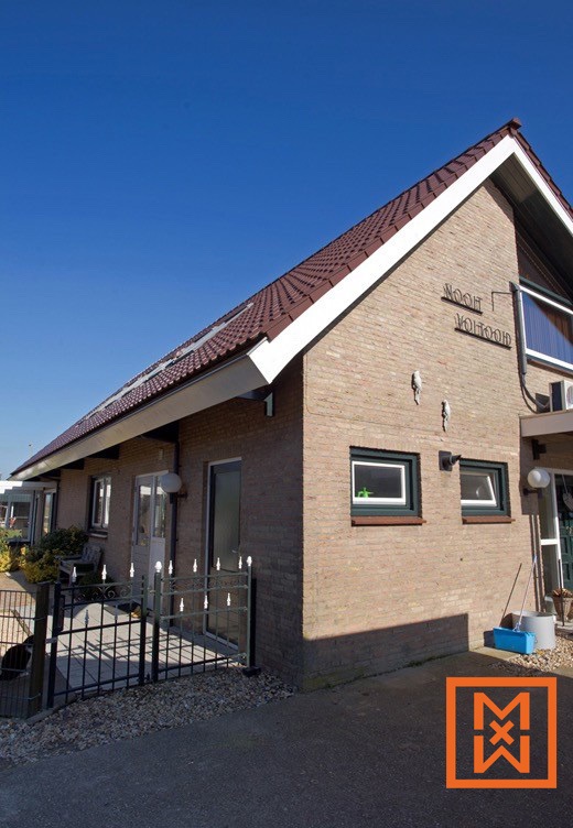 Een charmant bakstenen huis, "Noord Voornoud", heeft een roodbruin pannendak en een strak, zwart metalen hek. Zonlicht verlicht de buitenkant van het gebouw en creëert een warme en uitnodigende sfeer. Aan de zijkant van het huis zijn twee kleine vogeltjes bevestigd.