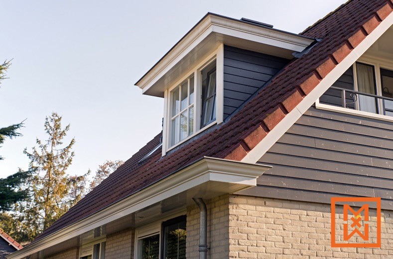 Close-up van een dakkapel met witte rand op een huis met donkergrijze gevelbekleding en een rood pannendak. De goten van het huis zijn schoon en goed onderhouden. Een deel van een bakstenen muur is zichtbaar.