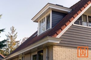 Een dakkapel met witte randen en meerdere ruiten voegt architectonische details toe aan het schuine dak van dit charmante huis. Het huis heeft donkergrijze gevelbekleding en een roodbruin pannendak, wat een opvallend contrast vormt met de helderblauwe lucht. Het tafereel straalt rust uit en suggereert een kwaliteitswoning.