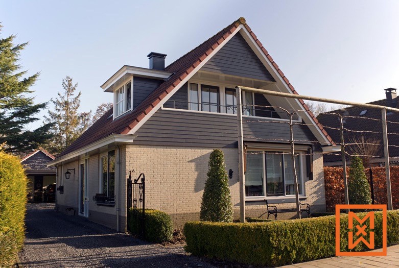 Charmante Nederlandse woning met grijze gevelbekleding en een beige bakstenen basis, een dakkapel en een goed onderhouden tuin. De woning heeft een rood pannendak en een kleine voortuin met netjes gesnoeide heggen.