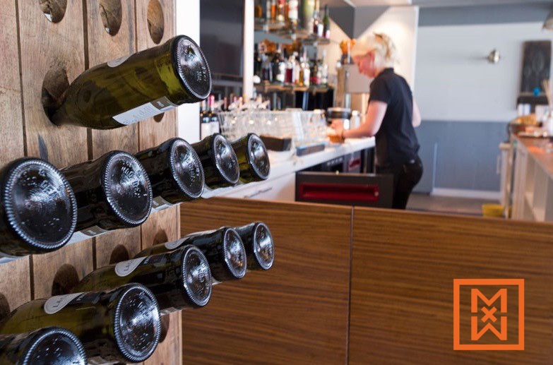 Close-up van een rustiek houten wijnrek met meerdere flessen wijn, schuin aflopend tegen een wazige achtergrond van een moderne bar met een barman aan het werk. De warme houttinten contrasteren met de donkergroene wijnflessen.