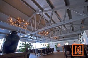 Ruim restaurantinterieur met hoge, grijs geschilderde plafonds met zichtbare houten balken. Twee kroonluchters met geweien hangen boven lange houten eettafels met donkere stoelen. Een grote donkere vaas staat op een bijzettafel bij een potplant. De algehele sfeer is verfijnd en rustiek.