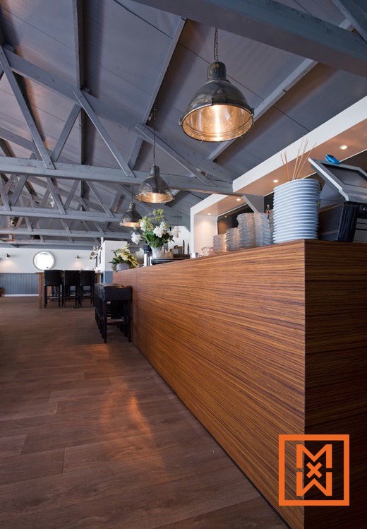 Modern restaurantinterieur met een lange, rijkelijk gekleurde houten bar en industriële hanglampen aan een hoog, grijs geschilderd balkenplafond. Donkere houten vloeren en eenvoudige zwarte barkrukken completeren het stijlvolle, minimalistische ontwerp.