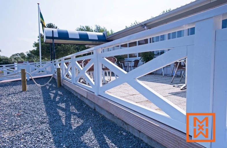Een wit houten hek met een X-patroon omringt een terras met metalen stoelen en tafels, met uitzicht op een gebouw met een gestreepte luifel en een vlaggenmast. Het terras is betegeld en het hek staat op een bruine houten basis. Het tafereel is licht en zonnig.
