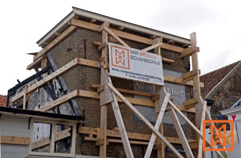 Een bakstenen gebouw, zwaar beschadigd en ondersteund door een uitgebreide houten steiger, ondergaat een structurele restauratie. Een spandoek van "Van Weenen Bouwbedrijf" met website en telefoonnummer is prominent aanwezig. Het gebouw vertoont sporen van brandschade.