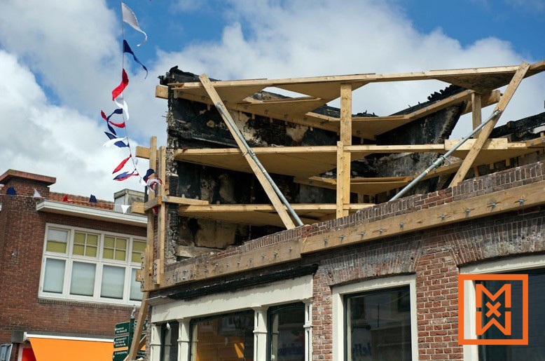 Een door brand beschadigd bakstenen gebouw wordt herbouwd. Houten steigers ondersteunen een tijdelijk dak boven de verkoolde resten van de bovenverdieping. Feestelijke slingers hangen ernaast, wat een schril contrast creëert tussen feestvreugde en verwoesting.