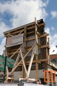 Een bakstenen gebouw dat een gevelrestauratie ondergaat, wordt ondersteund door een complex houten steigersysteem. Een spandoek van "Van Weenen Bouwbedrijf" is prominent aanwezig, met een reclame voor hun bouwactiviteiten. De zonnige dag en de deels bewolkte lucht vormen een heldere achtergrond voor dit stedelijke bouwtafereel.