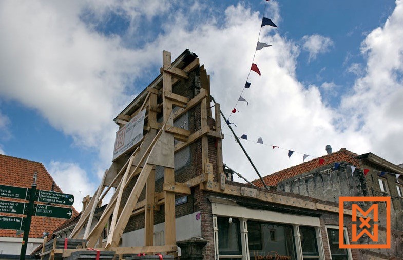 Een bakstenen gebouw ondergaat een gevelrestauratie, ondersteund door een complex houten geraamte. Het gebouw vertoont tekenen van ouderdom en schade, met steigers en beschermende bekleding. Kleurrijke slingers hangen boven de helderblauwe lucht.