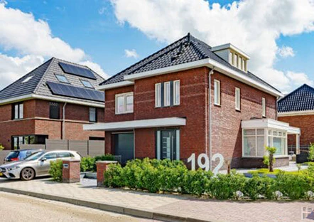 Modern bakstenen huis met nummer 192, strakke lijnen en groene beplanting in een rustige woonwijk. Zonnepanelen op het dak van het buurhuis.