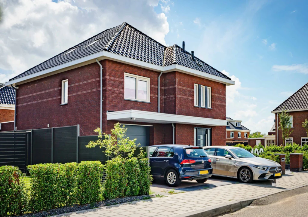 Modern bakstenen huis met donker dak, oprit met twee auto's (een blauwe en een zilveren). Groene haag en strakke lijnen geven een nette uitstraling. Blauwe lucht met wolken op de achtergrond.