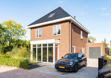 Modern bakstenen huis met donkerblauwe dakpannen en een oprit met een zwarte Mini Cooper. Een garage bevindt zich naast het huis, omgeven door groen.