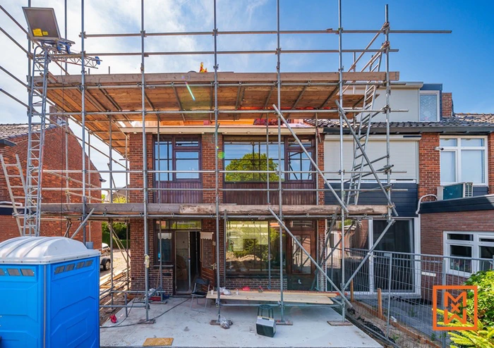 Scaffolding rondom bakstenen huis tijdens verbouwing met ramen en een blauw toilet op de voorgrond.