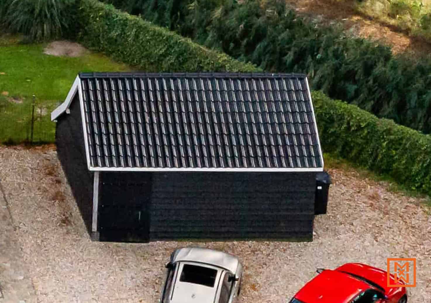 Zwarte schuur met pannendak tussen een grijze en rode auto, omgeven door groen struikgewas. Een rustieke uitstraling in een landelijke omgeving.