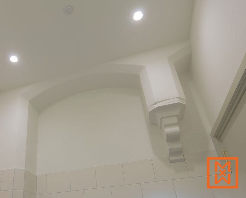 Close-up van een elegante, gebogen nis met inbouwverlichting, voorzien van een decoratieve, witte stucwerk console. De nis staat tegen een achtergrond van gebroken witte muren en lichtbeige vierkante tegels.