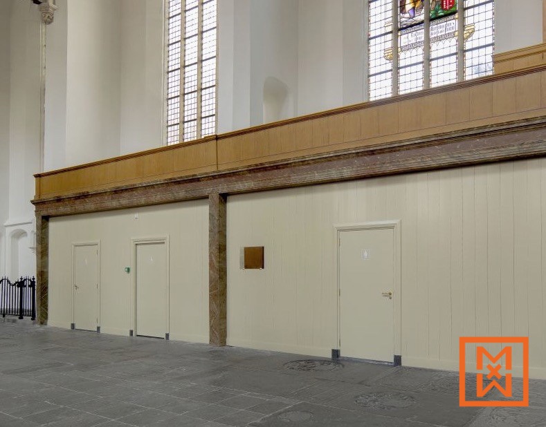Drie beige deuren met messing beslag zijn geplaatst in een lichtcrèmekleurige paneelmuur in een historische kerk. De muur is afgewerkt met een donkere afwerking van hout en marmer, onder een hoog, licht raam. De vloer is van donkergrijze steen.