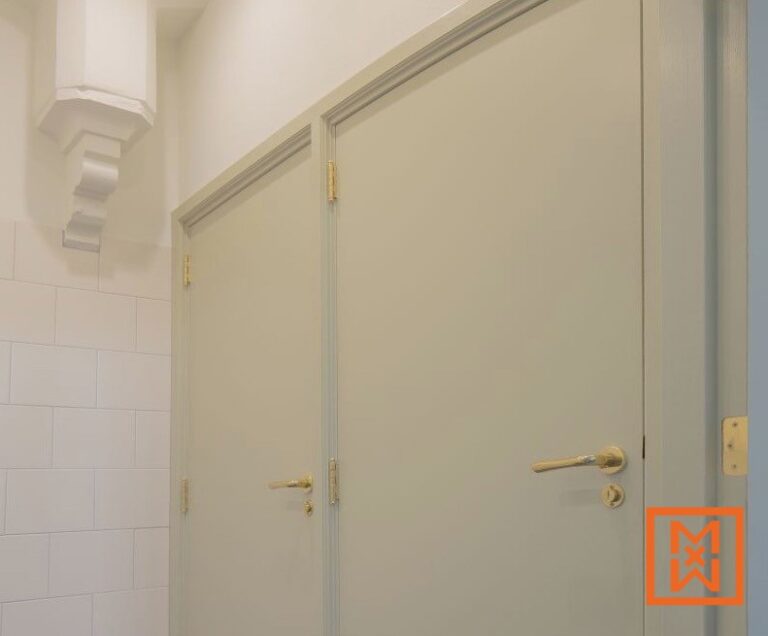 Twee lichtgroene binnendeuren met messing beslag zijn te zien in een lichte, moderne badkamer. De deuren hebben een subtiel gebogen bovenkant en staan tegen een witte muur met metrotegels. Een wit architectonisch detail is gedeeltelijk zichtbaar in de linkerbovenhoek.