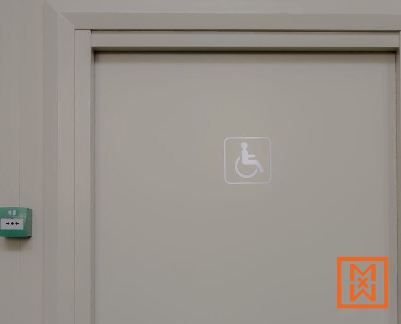 Close-up van een lichtbeige deur met een zilveren toegankelijkheidssymbool, wat aangeeft dat het een rolstoeltoegankelijk toilet betreft. Links op de muur is een groene noodknop zichtbaar.