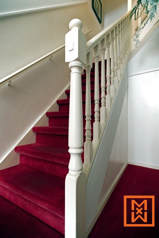 Elegante witte houten trap met een rode loper, gedraaide balustrades en een trappaal. De trap leidt naar de bovenverdieping in een goed verlicht interieur.