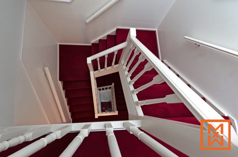 Bovenaanzicht van een wenteltrap met een dieprode loper en strakwitte leuningen. De trap loopt sierlijk rond en vormt een visueel aantrekkelijk designelement.
