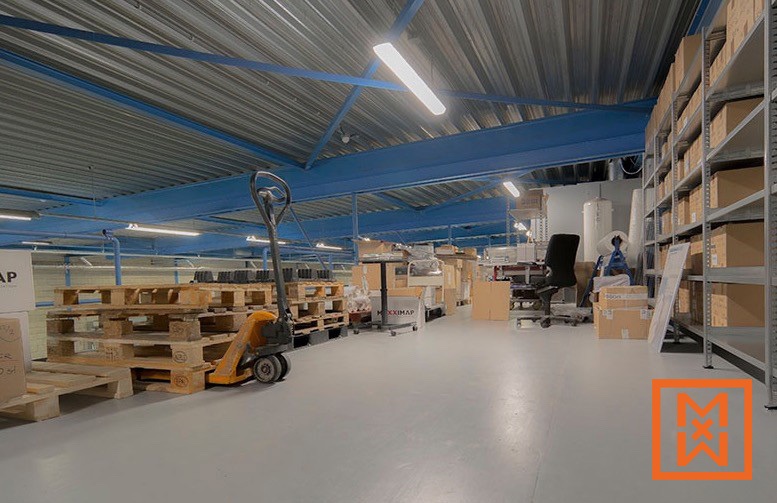 Een ruim magazijninterieur met een palletwagen naast stapels houten pallets en stellingen vol dozen. Het hoge plafond heeft zichtbare blauwe stalen balken en moderne ledverlichting. De algehele indruk is er een van efficiënte opslag en organisatie.