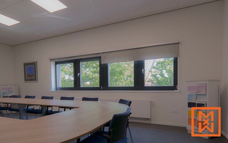 Moderne vergaderruimte met grote ramen, lichtgrijze rolgordijnen, een U-vormige vergadertafel van licht hout met donkerblauwe stoelen en een displaystandaard met kleurenstalen.