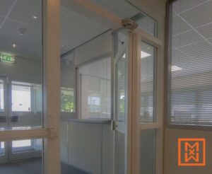 Modern kantoorinterieur met een strakke, witte receptiebalie en glazen wanden. Een halfopen deur met geïntegreerde jaloezieën geeft een lichte, luchtige werkruimte weer. Op de achtergrond is een bordje met de uitgang zichtbaar.