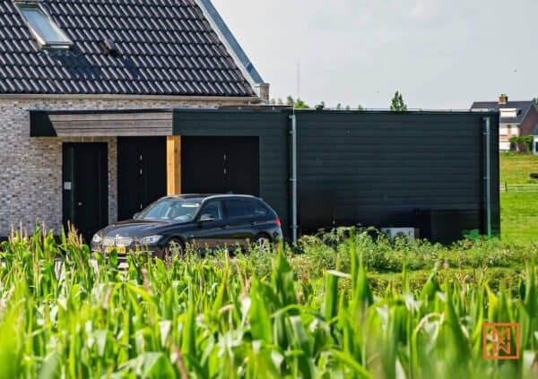 Zwarte BMW stationwagen geparkeerd voor een modern huis met donkere houten garage, omgeven door een veld met mais. Strakke architectuur en landelijk wonen.