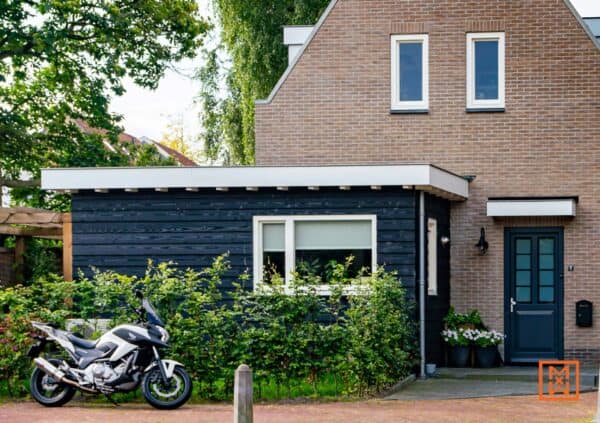 Een grijze Honda NC750X motorfiets geparkeerd voor een charmant huis met een blauwe deur en een donker houten aanbouw. Groene struiken sieren de voortuin.