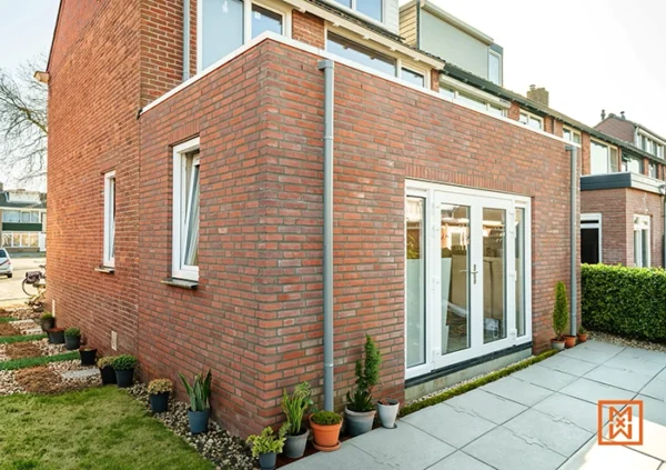 Uitbouw met rode bakstenen en witte kozijnen, met plantenbakken langs de muur.