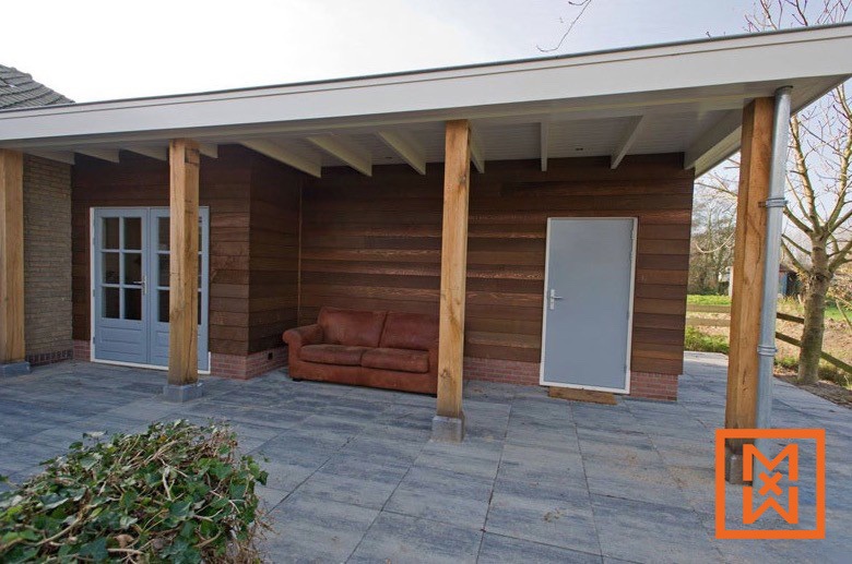 Rustieke houten pergola met een terras van grijze steen, een leren bank en een grijze buitendeur. De constructie heeft een donkerbruine houten buitenkant en is verbonden met een bakstenen huis.