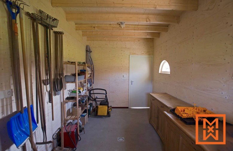 Een goed georganiseerde tuinschuur met houten wanden en zichtbare balken, waarin tuingereedschap, een grasmaaier en planken vol benodigdheden netjes zijn uitgestald. De ruimte is voorzien van een werkbank met een houten kast en een klein raam, wat een lichte en functionele werkplek creëert.