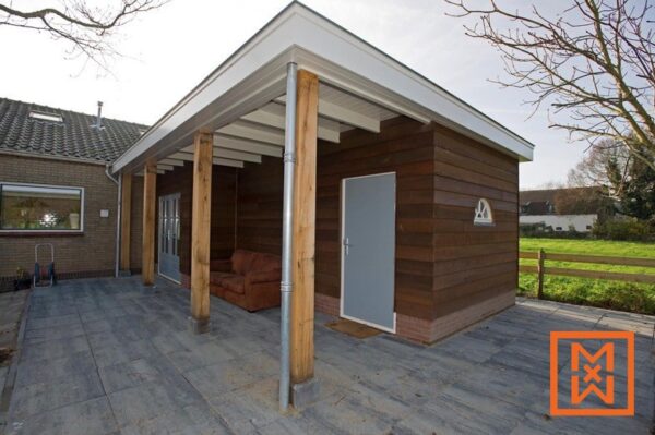 Moderne aanbouw aan de tuinkamer met donkerbruine houten gevelbekleding, een grijze deur en een overdekt terras met houten steunbalken en een terras van grijze steen. De constructie is verbonden met een bakstenen huis.