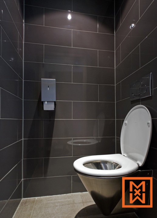 Modern, roestvrijstalen wandcloset in een klein, donkergrijs betegeld toilet. Een zeepdispenser en toiletspoeler zijn zichtbaar. Het minimalistische ontwerp zorgt voor een strakke en strakke uitstraling.