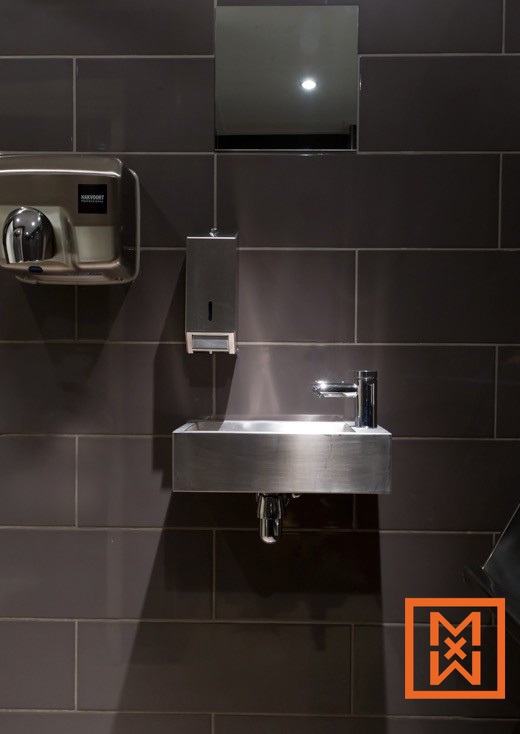 Strakke, roestvrijstalen wastafel met moderne kraan, handdroger en zeepdispenser, gemonteerd op een donkergrijze betegelde muur. Het minimalistische ontwerp creëert een strakke, eigentijdse esthetiek.