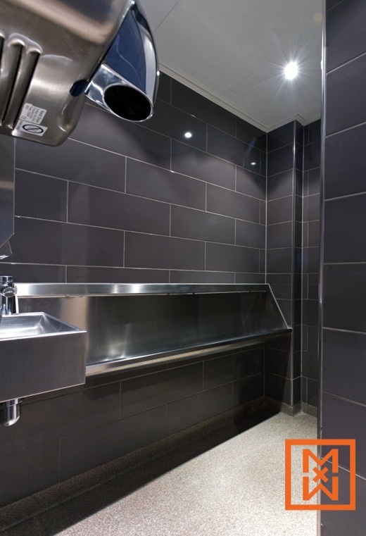 Modern roestvrijstalen urinoir en wastafel in een strak, donkergrijs betegeld herentoilet. Erboven hangt een metalen handdroger. De vloer is grijs gespikkeld.