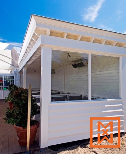 Witte buiteneethoek met glazen ramen en grijze, beklede zitkussens. De buitenkant is wit met planken bekleed en heeft een decoratieve witte rand. Op het terras ernaast staan planten in terracotta potten.