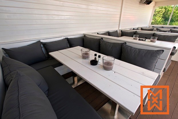 Moderne terrasstoelen met witte houten tafels en donkergrijze kussens. Het minimalistische ontwerp heeft een witte lattenwand als achtergrond. Kaarsen en peper- en zoutstelletjes staan op één tafel.