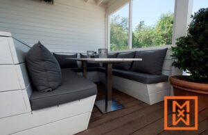 Moderne patio met een ingebouwde bank en grijze kussens rond een kleine vierkante tafel. De setting is licht en luchtig, met uitzicht op een weelderig groen landschap door de grote ramen.