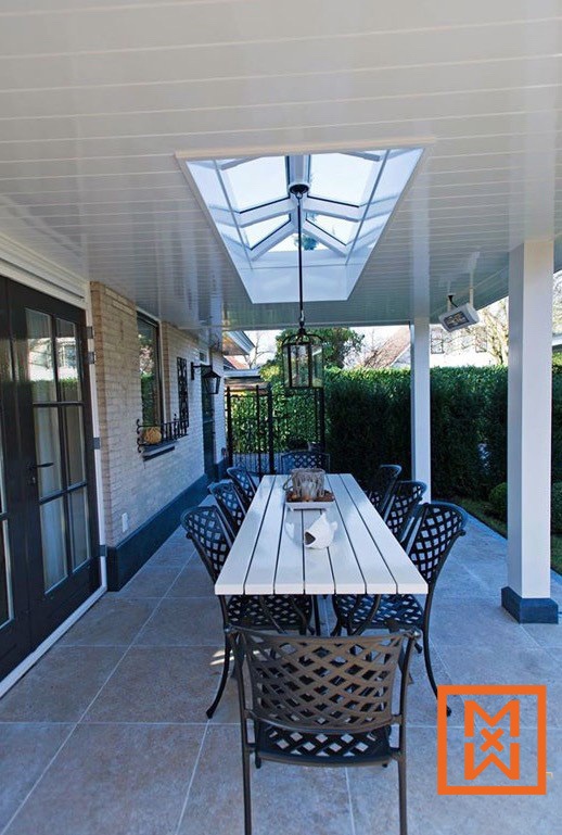 Een witte terrastafel met zwarte metalen stoelen staat op een lichtbeige tegelterras onder een wit paneelplafond met een dakraam. De setting is sereen en uitnodigend, perfect om buiten te eten. Een weelderige groene haag vormt de achtergrond.