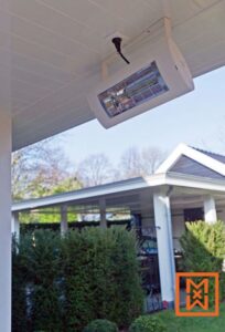 Witte elektrische terrasverwarmer gemonteerd onder een wit plafond, die warmte geeft op een terras met uitzicht op een weelderige groene haag en een modern huis.