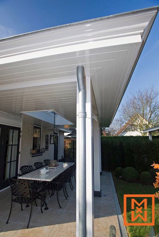 Modern terras met een witte pergola, beige tegelvloer en een lange witte eettafel met zwarte metalen stoelen. Een gegalvaniseerde regenpijp is zichtbaar op een witte steunbalk. Weelderig groen en een haag op de achtergrond.
