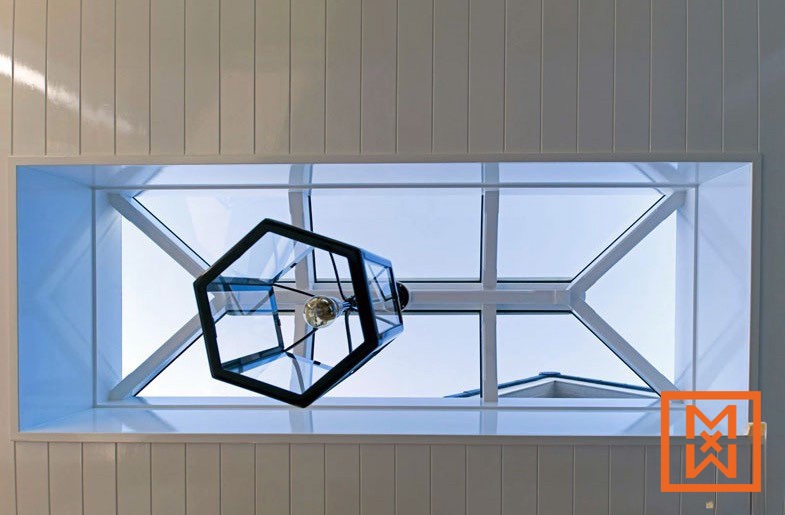 Een moderne zeshoekige glazen hanglamp hangt onder een stijlvol dakraam en verlicht een kamer met witte wanden van rabatdelen. Het dakraam heeft een strak wit frame en biedt uitzicht op de helderblauwe lucht.