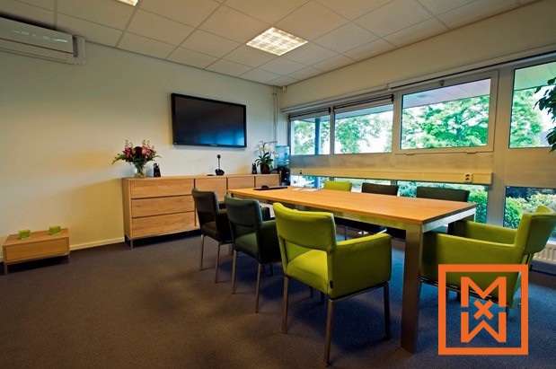 De moderne, lichte vergaderruimte is voorzien van een lichthouten vergadertafel, omringd door groene en donkergrijze bureaustoelen. Een flatscreen-tv en een lichthouten kast zijn zichtbaar, wat een professionele en uitnodigende sfeer creëert.