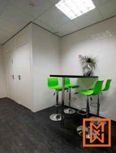Moderne kantoorkantine met een kleine zwarte tafel en drie felgroene barkrukken. De ruimte heeft witte tegelwanden en een donkere houtlook vloer. Op de achtergrond zijn twee witte deuren zichtbaar.